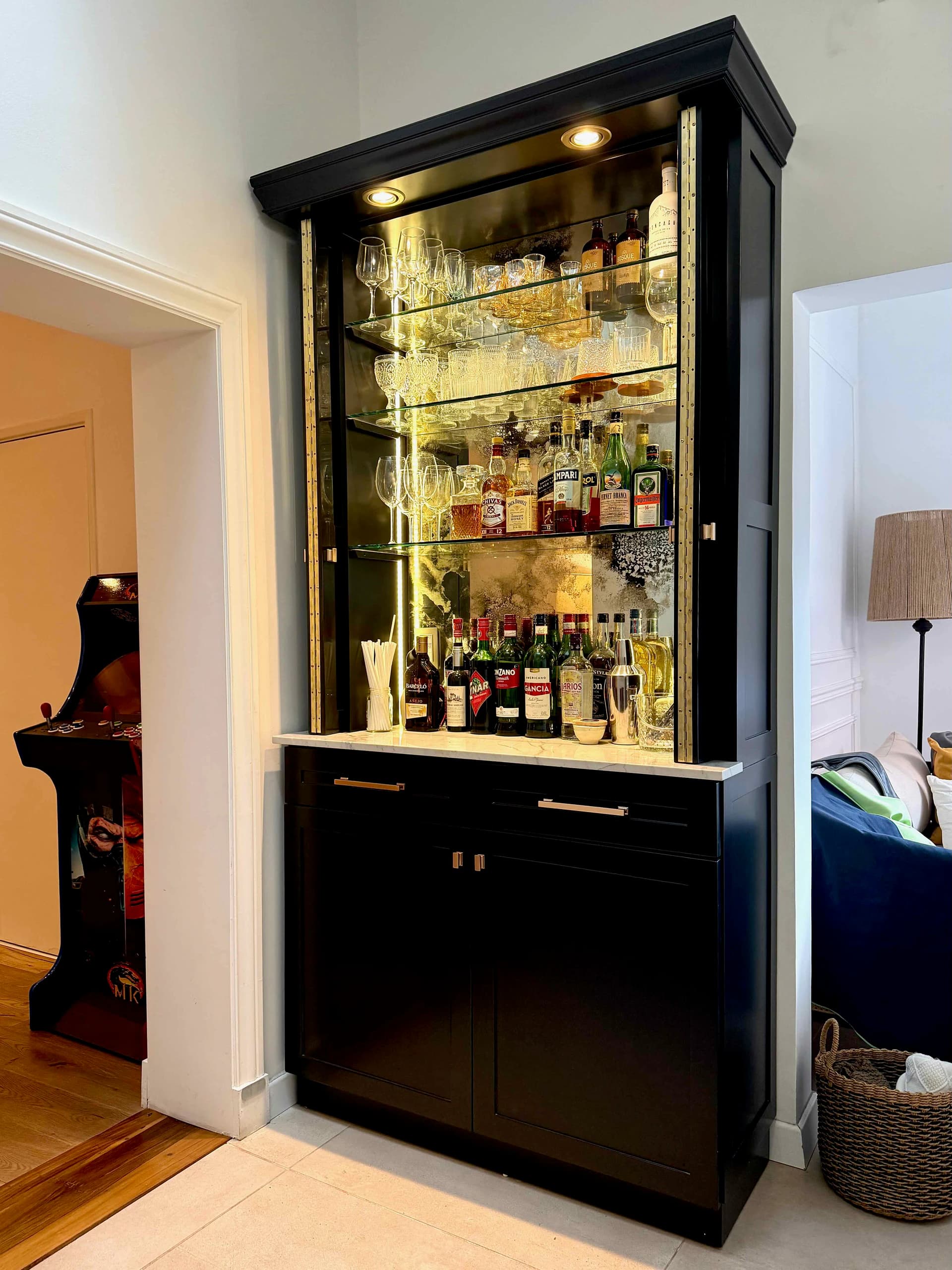 Mini bar oscuro abierto, con copas y botellas en los estantes iluminados y puertas en el guardado inferior.