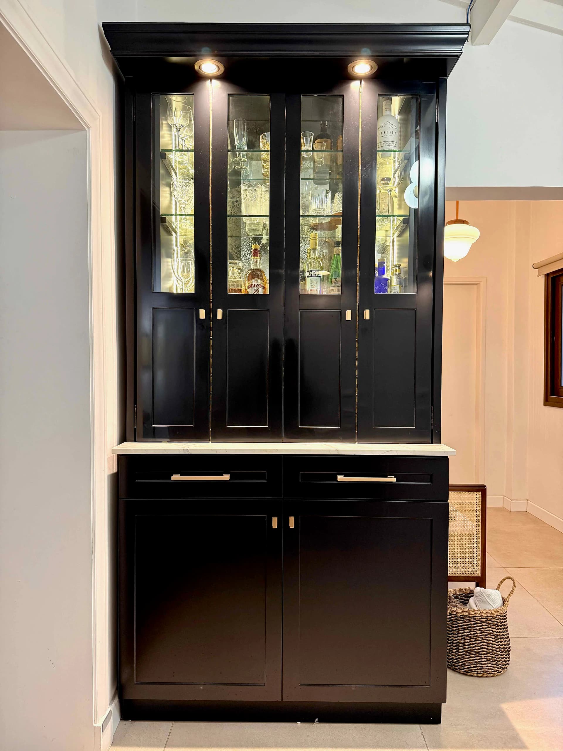 Mini bar con las puertas superiores cerradas, paños transparentes que dejan ver las copas y guardado inferior oscuro.